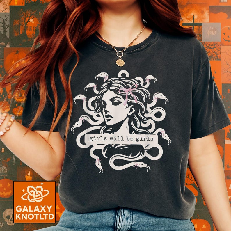 Puede incluir: Camiseta gris oscuro con una ilustraci&oacute;n blanca de una mujer con serpientes como cabello, y el texto "girls will be girls". El dise&ntilde;o incluye acentos rosas. Una prenda casual.