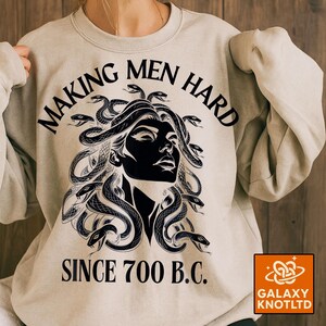 May include: A light beige sweatshirt with a black graphic of Medusa and snakes. The text "MAKING MEN HARD SINCE 700 B.C." is printed above and below the graphic. The Galaxy Knotltd logo is in the bottom right corner.