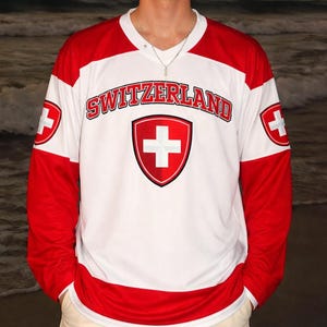Może przedstawiać: Biało-czerwona koszulka z długim rękawem z napisem "SWITZERLAND" na piersi. Koszulka posiada szwajcarską flagę na piersi i rękawach. Osoba ma na sobie szorty w kolorze khaki.