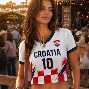 May include: A white crop top with navy blue trim, displaying "CROATIA" in navy blue and the number "10". The top features the Croatian coat of arms and red and blue accents. The person is wearing denim shorts and a brown belt.