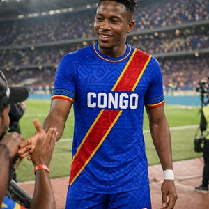 以下が含まれることがあります： 青色のサッカーユニフォームとショーツのセットで、白で「CONGO」の文字が入っています。ジャージには赤と黄色の斜めストライプとトリムがあります。ショーツには番号10が入っています。選手は笑顔で握手をしています。