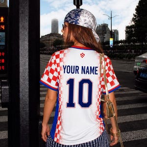 May include: A white and red checkered soccer jersey with the text "YOUR NAME" and the number "10" in blue. The jersey has blue accents and is worn with striped pants and a patterned headscarf. A gold purse hangs from the shoulder.
