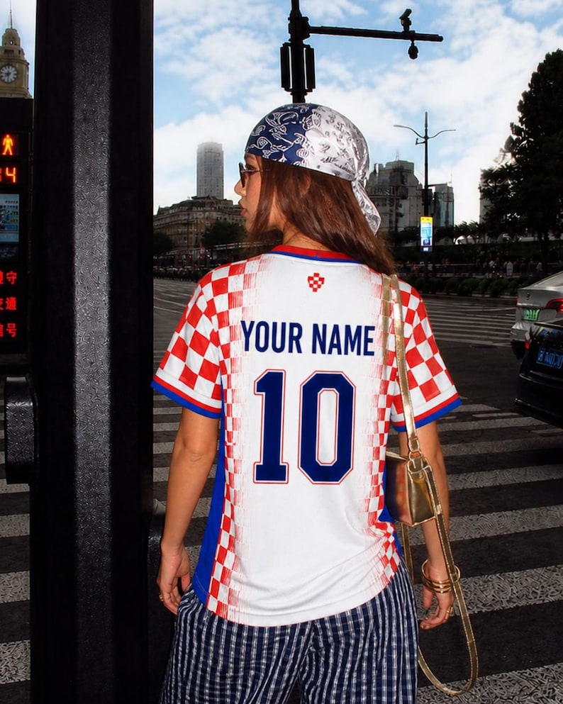 May include: A white and red checkered soccer jersey with the text "YOUR NAME" and the number "10" in blue. The jersey has blue accents and is worn with striped pants and a patterned headscarf. A gold purse hangs from the shoulder.