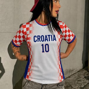 May include: A white and red checkered Croatia soccer jersey with the word "CROATIA" in blue and the number "10". The jersey has blue accents and is worn with a red baseball cap and gray denim jeans.