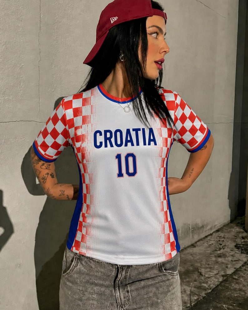May include: A white and red checkered Croatia soccer jersey with the word "CROATIA" in blue and the number "10". The jersey has blue accents and is worn with a red baseball cap and gray denim jeans.