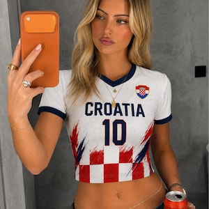 May include: White crop top with navy blue trim and the word "CROATIA" in navy blue. The number "10" is printed in navy blue. The top features a Croatian flag emblem and a red and white chequered pattern.