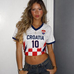 May include: White cropped football shirt with the word "CROATIA" and the number "10" in blue. The shirt features a red and white chequered pattern and a Croatian flag emblem. The shirt has blue trim and is worn with blue jeans.