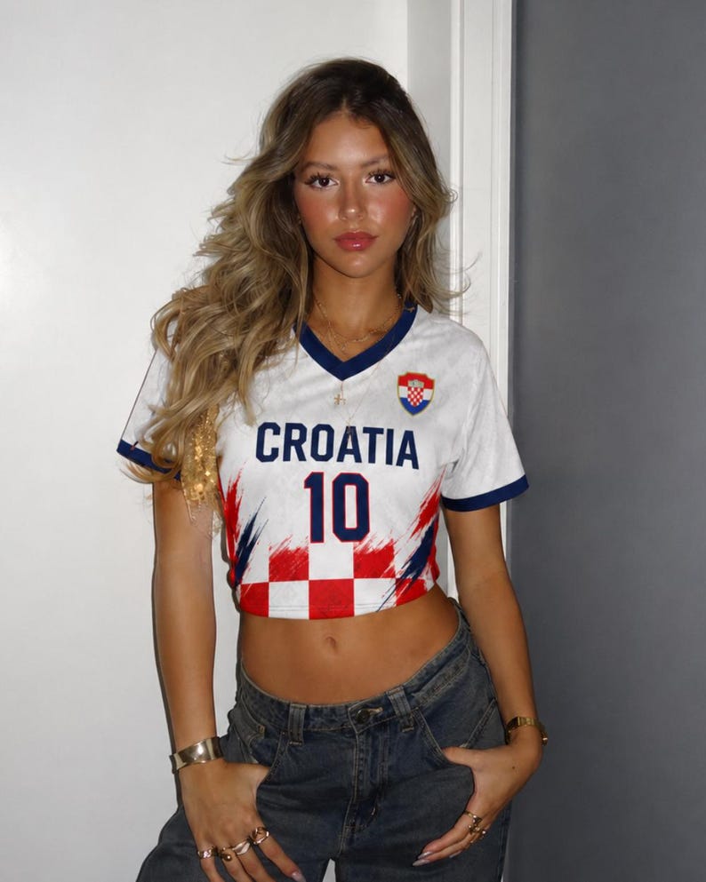May include: White cropped football shirt with the word "CROATIA" and the number "10" in blue. The shirt features a red and white chequered pattern and a Croatian flag emblem. The shirt has blue trim and is worn with blue jeans.