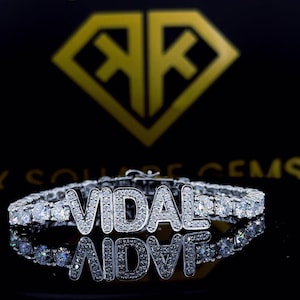 Peut inclure: Un bracelet de tennis en diamants avec le nom "VIDAL" épelé en lettres individuelles scintillantes. Le bracelet est posé sur un fond noir avec un logo doré.