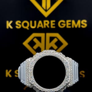 May include: A silver watch bezel encrusted with sparkling white gemstones. The bezel has a circular opening in the center and is set against a black background with the text "K SQUARE GEMS" and a logo.