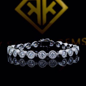 Peut inclure: Un bracelet en argent orné de plusieurs pierres précieuses rondes et transparentes. Chaque pierre est entourée de plus petites pierres transparentes. Le bracelet est présenté sur une surface noire réfléchissante, avec un logo doré en arrière-plan.