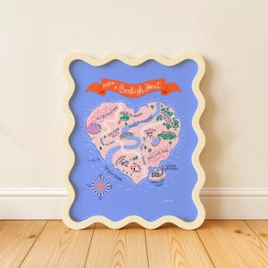 May include: A framed map titled "Map of a Bookish Heart" with a heart-shaped island design. The map features whimsical illustrations of mountains, rivers, and landmarks in shades of pink, green, and blue, set against a light blue background.