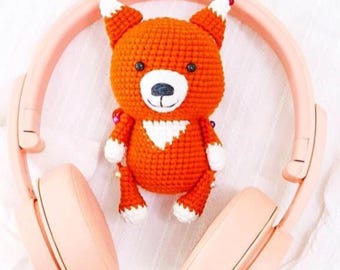May include: An orange crocheted fox toy with white accents and a white chest sits between a pair of pink wireless headphones. The headphones are folded and resting on a white surface.