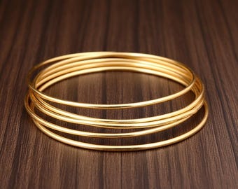 May include: A stack of gold-colored bangles. The bangles are thin and round, with a smooth, polished surface. They are arranged in a slightly overlapping pattern on a dark brown wooden surface.