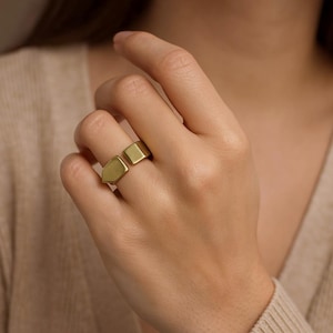 Op de afbeelding: Een goudkleurige ring met een geometrisch ontwerp. De ring heeft een vierkant en een schildvormig element, waardoor een open ontwerp ontstaat. De ring wordt aan een vinger gedragen en toont zijn moderne en minimalistische stijl.