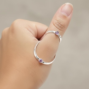 Könnte beinhalten: Ein silberner Ring mit zwei lila Edelsteinen. Der Ring ist für den Daumen konzipiert und umschließt den Finger. Die Edelsteine sind klein und rund.