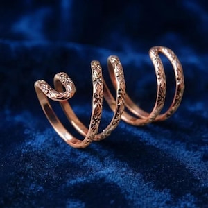 Pode incluir: Um anel de metal em tom de ouro rosa com um design em espiral de várias voltas. A superfície da banda é decorada com um padrão repetitivo em relevo de flores ou cruzes ao longo de todo o seu comprimento, criando um aspeto envolvente.