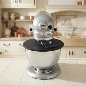 Könnte beinhalten: Eine silberne KitchenAid Artisan Küchenmaschine mit metallischem Finish, bestehend aus einem abgerundeten Kopf, einer Edelstahlschüssel und einem schwarzen, kreisförmigen Spritzschutz, der oben auf der Schüssel befestigt ist. Der Markenname steht vorne.