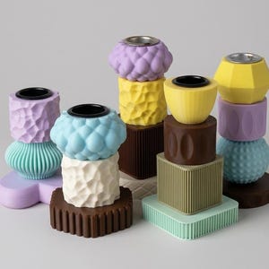 Könnte beinhalten: Eine Sammlung modularer Kerzenhalter in verschiedenen geometrischen Formen und Pastellfarben wie Lavendel, Hellblau, Gelb, Minzgrün und Braun. Jedes Teil weist unterschiedliche Texturen wie Rillen, Noppen und glatte Oberflächen auf.