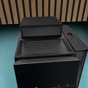 May include: A modern, black coffee machine with a rectangular design. The machine features a top-mounted box, a tray with horizontal grooves, and a control panel with icons. The overall aesthetic is sleek and minimalist.