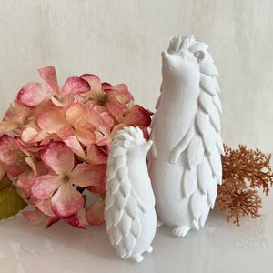 Peut inclure: Deux figurines de hérissons blanches debout, l'une plus grande que l'autre, placées à côté d'un bouquet d'hortensias roses et blancs et d'un petit brin de feuillage brun séché, le tout présentant une texture de surface mate et lisse.