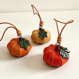 Puede incluir: Tres colgantes de calabaza de cuero en naranja, bronceado claro y rojo, cada uno con una hoja verde y un lazo de cuero. Los colgantes miden aproximadamente 5 cm por 5 cm. El texto en la imagen dice: Size: approx. 1.97" x 1.97" (5x5 cm).