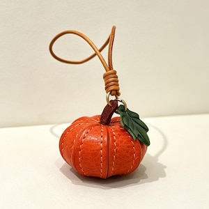 May include: A small, orange, pumpkin-shaped charm crafted with visible stitching. It features a dark brown stem, a single green leaf, and a thin, looped tan leather cord attached to the top for hanging or attachment to other items.