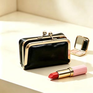 Puede incluir: Un estuche rectangular negro con un cierre de marco metálico dorado tipo boquilla. Junto al estuche hay un labial rojo con tapa rosa claro y un pequeño espejo compacto dorado abierto que contiene dos tonos de polvo compacto.