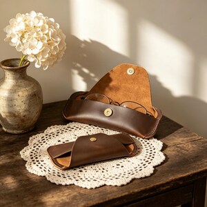 May include: Two dark brown leather glasses cases with visible stitching and gold-toned metal snap closures. One case is open, revealing a pair of thin-framed eyeglasses inside, while the other case is closed and positioned in the foreground.