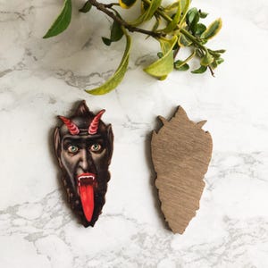 May include: Wooden cutout of a devil's face with red horns, green eyes, and a long red tongue. Next to it is a wooden cutout of the same shape, but unpainted. The cutouts are on a white and gray marble surface.