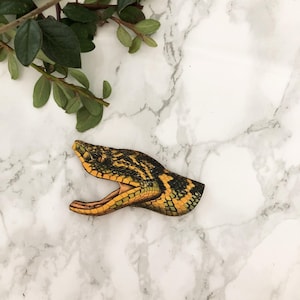Puede incluir: Broche de madera de serpiente con la boca abierta, con un patrón detallado de escamas amarillas, negras y verdes. El broche está sobre un fondo de mármol blanco con follaje verde.