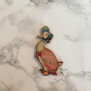 Puede incluir: Figura de pato de madera con un gorro azul y un vestido estampado en tonos rojos, rosas y verdes. El pato tiene pico y patas naranjas. La figura está sobre un fondo de mármol blanco y gris.