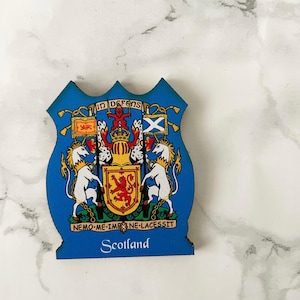 Pode incluir: Um íman azul em forma de escudo com o brasão escocês. Apresenta um leão vermelho rampante, unicórnios brancos e a palavra "Scotland". O íman tem uma borda decorativa e é provavelmente uma lembrança.