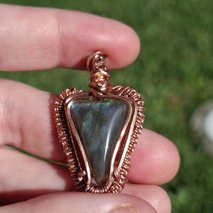 May include: A triangular labradorite gemstone pendant encased in intricate copper wire wrapping. The stone displays a dark grey and blue iridescent sheen, secured by a coiled copper bail at the top, creating a detailed metalwork frame around the stone.
