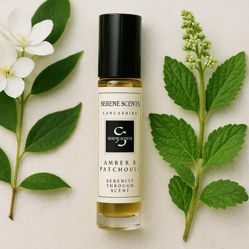 May include: A small amber-colored essential oil roller bottle with a black cap. The label reads "SERENE SCENTS" and "AMBER & PATCHOULI". The bottle is flanked by white flowers and green leaves, creating a natural, spa-like aesthetic.