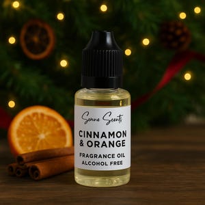 May include: A clear glass bottle with a black cap, labelled "Cinnamon & Orange Fragrance Oil Alcohol Free." The bottle is next to cinnamon sticks and an orange slice. The background includes a Christmas tree and lights.