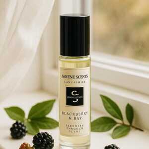 May include: A perfume bottle with a black cap and a label that reads "SERENE SCENTS LANCASHIRE" and "BLACKBERRY & BAY". The bottle is surrounded by blackberries and green leaves. The scent is described as "SERENITY THROUGH SCENT".