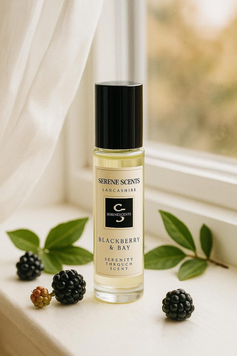 May include: A perfume bottle with a black cap and a label that reads "SERENE SCENTS LANCASHIRE" and "BLACKBERRY & BAY". The bottle is surrounded by blackberries and green leaves. The scent is described as "SERENITY THROUGH SCENT".