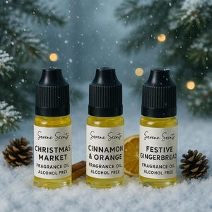 May include: Three small glass bottles of fragrance oil with black caps, labelled "Christmas Market," "Cinnamon & Orange," and "Festive Gingerbread." The bottles contain yellow liquid and are on a snowy surface with pine cones and an orange slice.