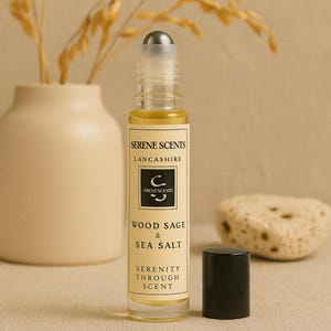 May include: A clear glass roll-on bottle with a silver cap, filled with a yellow liquid. The label reads "Serene Scents Lancashire" and "Wood Sage & Sea Salt". A black cap sits beside the bottle. The background includes a vase and decorative stone.
