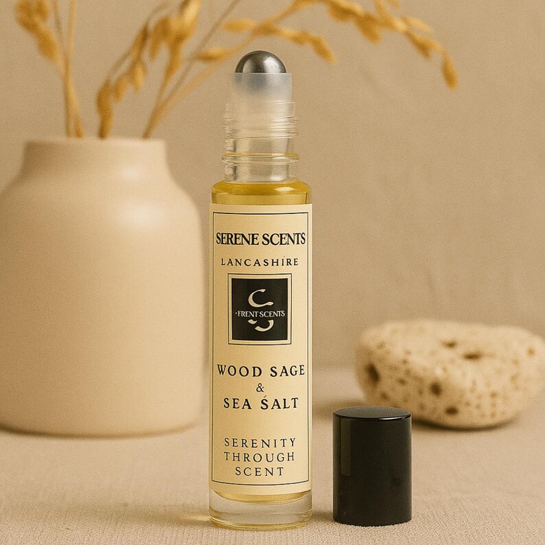 May include: A clear glass roll-on bottle with a silver cap, filled with a yellow liquid. The label reads "Serene Scents Lancashire" and "Wood Sage & Sea Salt". A black cap sits beside the bottle. The background includes a vase and decorative stone.