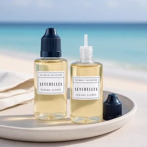 May include: Two clear glass bottles of "Seychelles Serene Scents" perfume from The White Collection. The bottles have white labels with black text and dark blue caps. One cap is off. The bottles sit on a white plate with a blurred ocean background.