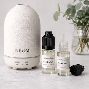 May include: A white essential oil diffuser with the brand name "NEOM" printed on the front. Two small glass bottles of "Serene Scents Tranquility" premium fragrance oil are next to the diffuser. The diffuser has "LIGHT" and "MIST" buttons.