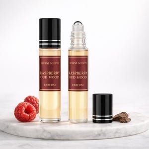 May include: Two perfume bottles with black caps and labels reading "Serene Scents Raspberry Oud Mood Parfum." One bottle has a rollerball applicator. Fresh raspberries are placed nearby. The bottles are on a white marble surface.