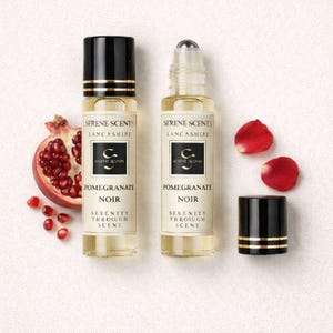 May include: Two perfume bottles with black and gold caps, labeled "Pomegranate Noir" by Serene Scents. A halved pomegranate and red petals are arranged nearby. The bottles have a roll-on applicator.
