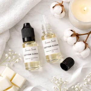 May include: Two small glass bottles of "Cotton Clouds" premium fragrance oil with white labels, a black cap, and a white dropper. The bottles are next to a white towel, cotton bolls, a lit candle, and wax melts. The overall theme is relaxation and aromatherapy.