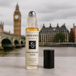 May include: A clear glass roll-on bottle with a silver cap and a black cap next to it. The label reads "Serene Scents Lancashire" and "Pomegranate Noir". The background features a blurred view of a city with a clock tower.