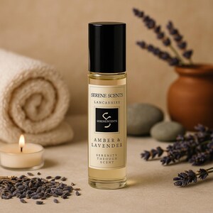 May include: A clear glass bottle with a black cap, labeled "Serene Scents Lancashire" and "Amber & Lavender." The bottle is surrounded by a rolled towel, a lit candle, lavender buds, and a small vase with lavender sprigs. The overall color palette is neutral with warm tones.