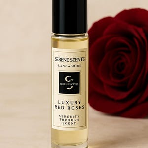 May include: A clear glass perfume bottle with a black cap, labeled "Serene Scents Lancashire" and "Luxury Red Roses." A deep red rose is in the background. The bottle contains a light yellow liquid. The label also says "Serenity Through Scent."
