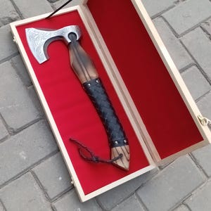 May include: A Viking-style axe in a wooden presentation box. The axe head is silver with etched designs, and the handle is wrapped in black leather. The box has a red velvet lining and a wooden frame.